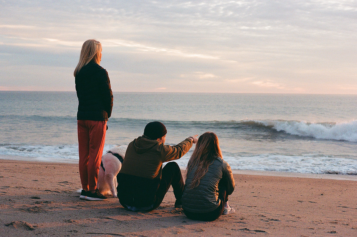 Three people and a dog on a sandy beach watching the waves at sunset